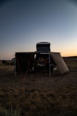 A campsite set up in an open field during the early evening. A vehicle with a rooftop tent and an attached canopy is in the center. The sky is clear with a visible crescent moon, and the surrounding landscape is mostly flat with sparse vegetation.