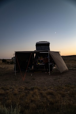 A campsite set up in an open field during the early evening. A vehicle with a rooftop tent and an attached canopy is in the center. The sky is clear with a visible crescent moon, and the surrounding landscape is mostly flat with sparse vegetation.