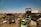 A sprawling junkyard filled with various types of scrap metal, including old vehicles, tires, and barrels scattered across dry, grassy terrain under a clear blue sky. Rusted car frames and deteriorating machinery are prominent among the debris.