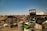 A sprawling junkyard filled with various types of scrap metal, including old vehicles, tires, and barrels scattered across dry, grassy terrain under a clear blue sky. Rusted car frames and deteriorating machinery are prominent among the debris.