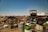 A sprawling junkyard filled with various types of scrap metal, including old vehicles, tires, and barrels scattered across dry, grassy terrain under a clear blue sky. Rusted car frames and deteriorating machinery are prominent among the debris.