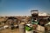 A sprawling junkyard filled with various types of scrap metal, including old vehicles, tires, and barrels scattered across dry, grassy terrain under a clear blue sky. Rusted car frames and deteriorating machinery are prominent among the debris.