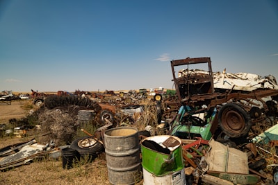 A sprawling junkyard filled with various types of scrap metal, including old vehicles, tires, and barrels scattered across dry, grassy terrain under a clear blue sky. Rusted car frames and deteriorating machinery are prominent among the debris.