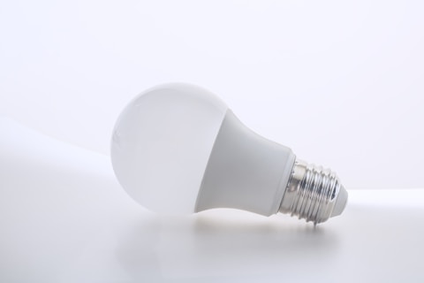 A white LED bulb lies on its side against a plain, light-colored background. The metallic base near the end of the bulb contrasts slightly against the smooth, soft texture of the bulb's surface.