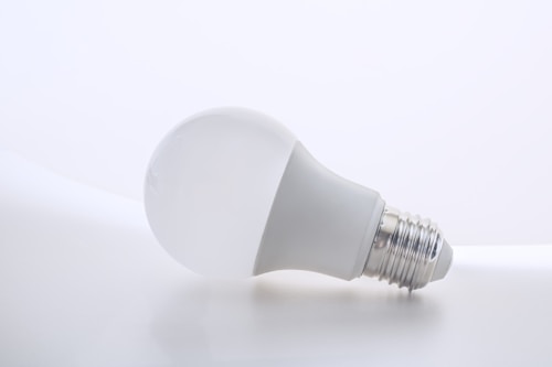 A white LED bulb lies on its side against a plain, light-colored background. The metallic base near the end of the bulb contrasts slightly against the smooth, soft texture of the bulb's surface.