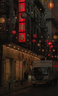 A dimly lit urban street scene features a large red vertical sign with Chinese characters, surrounded by hanging lanterns in shades of red, orange, and white. A white truck with graffiti is parked alongside the street, next to a bicycle. A person stands near the entrance of a building, holding a cup.