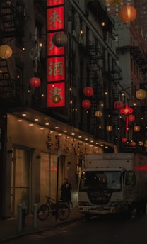 A dimly lit urban street scene features a large red vertical sign with Chinese characters, surrounded by hanging lanterns in shades of red, orange, and white. A white truck with graffiti is parked alongside the street, next to a bicycle. A person stands near the entrance of a building, holding a cup.
