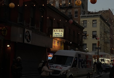 A street scene during dusk or early evening, featuring buildings adorned with string lights and colorful hanging lanterns. There is a FedEx delivery van parked on the street and a wine and liquor store with a lighted sign. Several people are walking along the sidewalk, bundled up for colder weather.