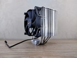 A large cooling fan attached to a metallic heatsink assembly rests on a wooden surface. The design features numerous metal fins and pipes extending down, reflecting light. A black cable is visible, suggesting connectivity for power or electronics.