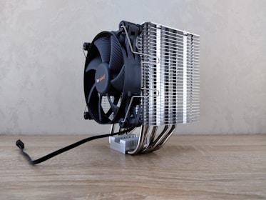 A large cooling fan attached to a metallic heatsink assembly rests on a wooden surface. The design features numerous metal fins and pipes extending down, reflecting light. A black cable is visible, suggesting connectivity for power or electronics.