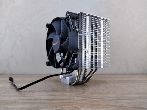 A large cooling fan attached to a metallic heatsink assembly rests on a wooden surface. The design features numerous metal fins and pipes extending down, reflecting light. A black cable is visible, suggesting connectivity for power or electronics.