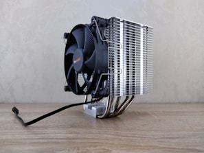 A large cooling fan attached to a metallic heatsink assembly rests on a wooden surface. The design features numerous metal fins and pipes extending down, reflecting light. A black cable is visible, suggesting connectivity for power or electronics.