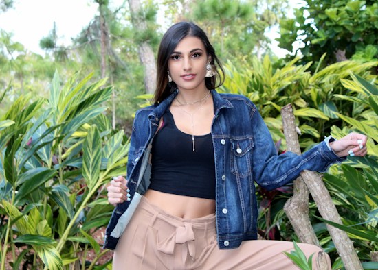 A person stands outdoors surrounded by greenery, wearing a denim jacket, black top, and beige pants. They pose confidently, resting one arm on a wooden fence and the other on their hip. The lush foliage provides a vibrant, natural backdrop, while the person's expression and attire add a touch of modern style.