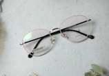 A pair of eyeglasses with pink frames and black temples rests on a light-colored textured surface. A barcode label is visible on one of the temples, and a spider logo is featured on the lens. Some foliage is partially visible in the corner.