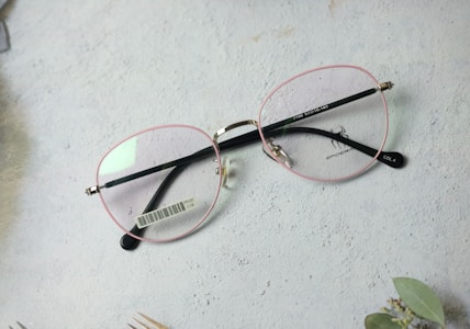 A pair of eyeglasses with pink frames and black temples rests on a light-colored textured surface. A barcode label is visible on one of the temples, and a spider logo is featured on the lens. Some foliage is partially visible in the corner.