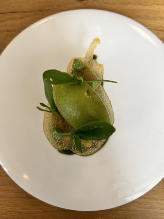 A neatly arranged dish featuring a green dessert topped with small green leaves and delicate pea-sized elements. The presentation is minimalist, placed on a simple white plate and set on a light wooden table.