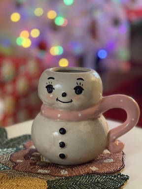 A ceramic mug shaped like a snowman with a pink scarf and big painted eyes. It has black buttons and a friendly smile. The mug sits on a textured, colorful coaster with a blurred background featuring colorful bokeh lights.