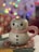 A ceramic mug shaped like a snowman with a pink scarf and big painted eyes. It has black buttons and a friendly smile. The mug sits on a textured, colorful coaster with a blurred background featuring colorful bokeh lights.