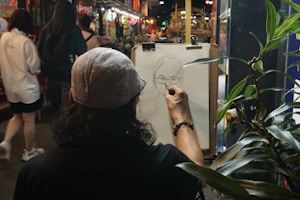 An artist is drawing a portrait of a person on a bustling street. The scene is vibrant with several people walking by, and the artist is focused on sketching the details of the person's face. The image captures a mix of natural elements with plants in the foreground, and a lively indoor market is visible in the background.