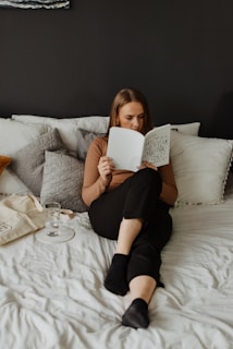 A woman is seated comfortably on a bed, engrossed in reading a book. The bed is adorned with various pillows, and a glass of water along with a tote bag is placed beside her. The room exudes a cozy and tranquil atmosphere, with muted colors adding to the serene setting.
