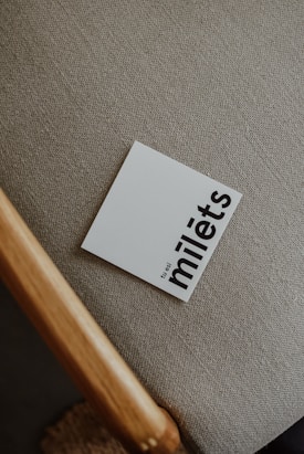 A card with bold black text sits on a textured, neutral-colored fabric. The card reads 'tu esi milēts' in lowercase letters. Part of a wooden chair is visible in the image.