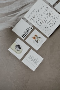 Several artfully arranged cards lie on a textured gray surface. One card features abstract black ink patterns with the word 'IDENTITATE', another displays a sunflower illustration, and others contain text with varying fonts, including cursive writing. A sheer white fabric is draped artistically near the cards.