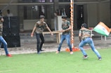 Several young individuals are actively engaging in an outdoor activity on a grass field. They are dressed in matching camouflage shirts and jeans. One person is holding the national flag of India. In the background, there is a building with a tiled pathway and a stack of large black speakers. Decorative orange and white garlands hang from a pole.