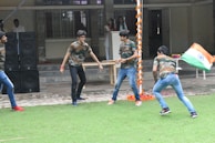 Several young individuals are actively engaging in an outdoor activity on a grass field. They are dressed in matching camouflage shirts and jeans. One person is holding the national flag of India. In the background, there is a building with a tiled pathway and a stack of large black speakers. Decorative orange and white garlands hang from a pole.