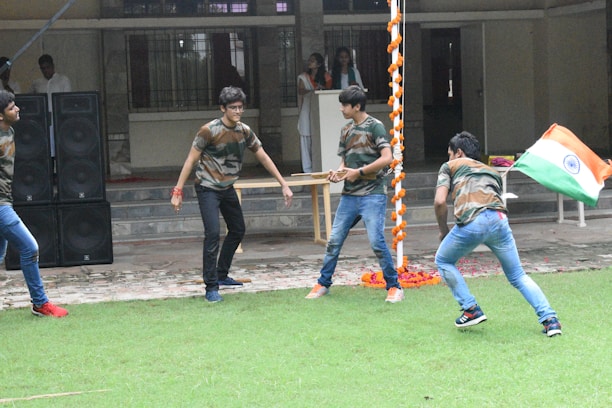 Several young individuals are actively engaging in an outdoor activity on a grass field. They are dressed in matching camouflage shirts and jeans. One person is holding the national flag of India. In the background, there is a building with a tiled pathway and a stack of large black speakers. Decorative orange and white garlands hang from a pole.