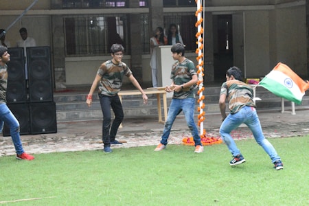 Several young individuals are actively engaging in an outdoor activity on a grass field. They are dressed in matching camouflage shirts and jeans. One person is holding the national flag of India. In the background, there is a building with a tiled pathway and a stack of large black speakers. Decorative orange and white garlands hang from a pole.
