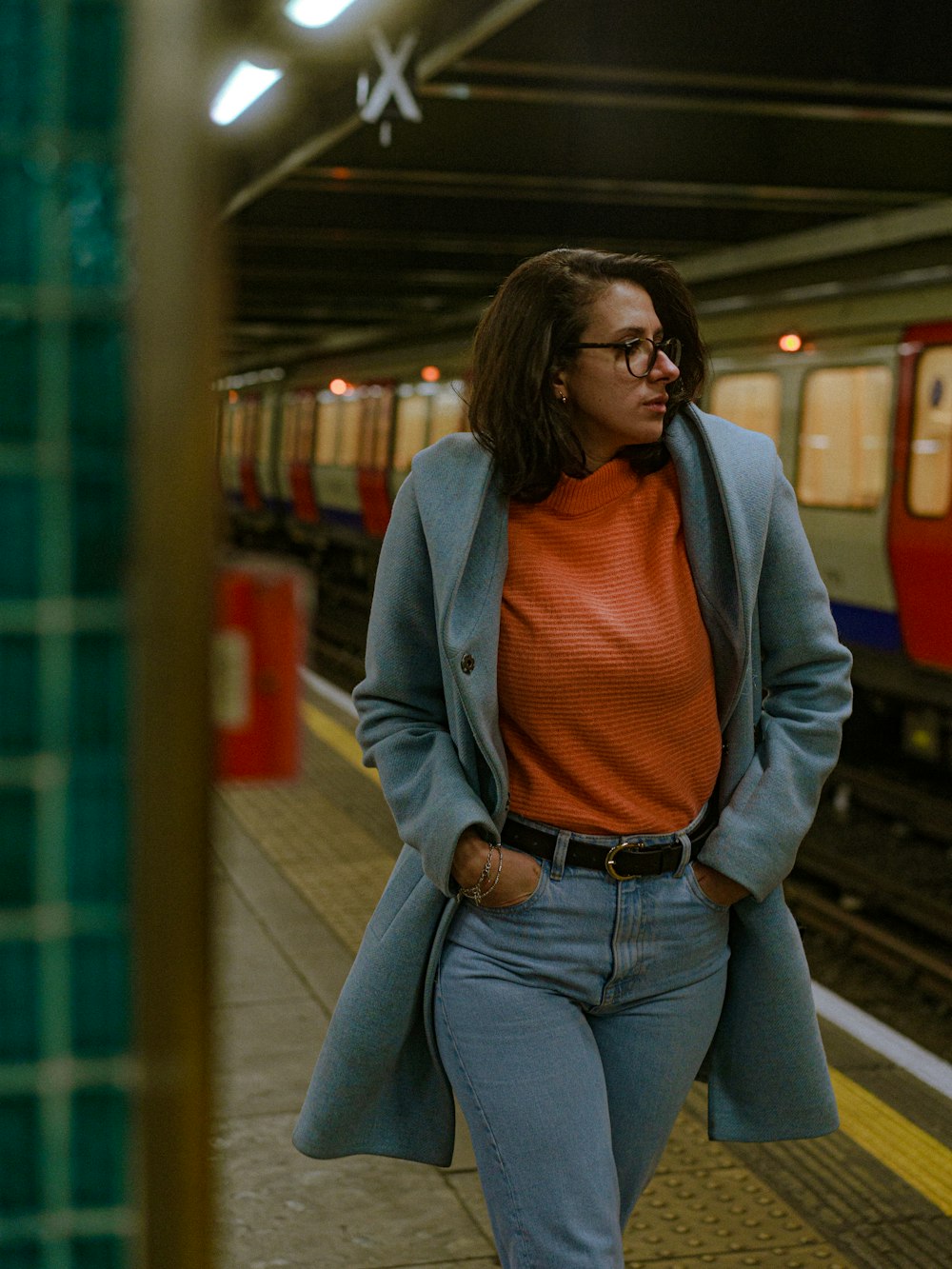 a woman in a blue coat and orange sweater is walking on a subway platform