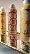 Three brightly colored, patterned aerosol cans are arranged in a line on a reflective surface. The cans have a retro design with floral and geometric motifs in shades of orange, yellow, pink, blue, and red. The central canister is labeled 'perk up dry shampoo' and accompanied by the words 'shampooing sec'. The brand 'amika:' is prominently displayed on each can.
