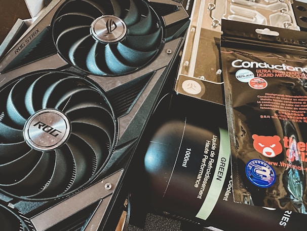 A close-up of a computer graphics card with two large cooling fans labeled ROG Strix, alongside a package of thermal paste labeled Conductonaut and a bottle of liquid cooling solution. The items appear to be for enhancing computer performance and cooling.