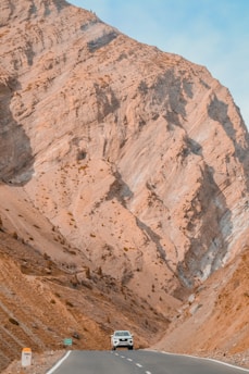 A white car drives along a winding road surrounded by towering, rugged, brown rocky mountains. The scene conveys a sense of isolation and adventure amidst vast natural landscapes.