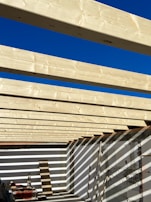 Several light-colored wooden beams are aligned parallel across an open-roof frame, casting shadows on the white walls below. The sky above is clear and bright blue, contributing to the high contrast with the wooden structure. Construction materials and tools are present on the floor.