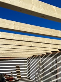 Several light-colored wooden beams are aligned parallel across an open-roof frame, casting shadows on the white walls below. The sky above is clear and bright blue, contributing to the high contrast with the wooden structure. Construction materials and tools are present on the floor.