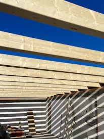 Several light-colored wooden beams are aligned parallel across an open-roof frame, casting shadows on the white walls below. The sky above is clear and bright blue, contributing to the high contrast with the wooden structure. Construction materials and tools are present on the floor.