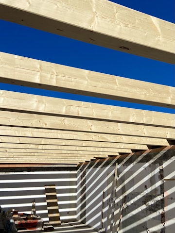 Several light-colored wooden beams are aligned parallel across an open-roof frame, casting shadows on the white walls below. The sky above is clear and bright blue, contributing to the high contrast with the wooden structure. Construction materials and tools are present on the floor.