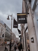 A bustling city street lined with elegant shops, several signs for a fashion brand prominently displayed. The architecture features ornate, historic buildings, and a crowd of people walks along the sidewalk, some carrying shopping bags. The sky is overcast, adding a subdued atmosphere to the scene.