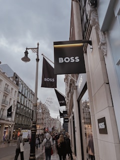 A bustling city street lined with elegant shops, several signs for a fashion brand prominently displayed. The architecture features ornate, historic buildings, and a crowd of people walks along the sidewalk, some carrying shopping bags. The sky is overcast, adding a subdued atmosphere to the scene.