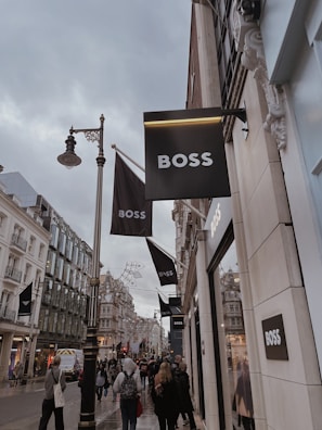 A bustling city street lined with elegant shops, several signs for a fashion brand prominently displayed. The architecture features ornate, historic buildings, and a crowd of people walks along the sidewalk, some carrying shopping bags. The sky is overcast, adding a subdued atmosphere to the scene.