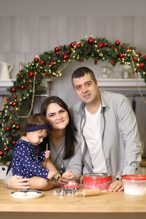 A family of three is gathered in a cozy kitchen setting, with a festive atmosphere created by a Christmas garland in the background. The parents smile at the camera, while the child, dressed in a polka-dotted outfit, concentrates on playing with a toy car. Baking items are laid out on the wooden table in front of them, including cookie cutters and a rolling pin.