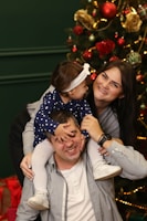 A family poses playfully in front of a decorated Christmas tree. The young child, wearing a headband and dotted outfit, is sitting on the man's shoulders. The woman beside them is smiling joyfully, while holding the child securely. Gift boxes wrapped in red paper are visible in the background.