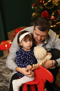 A young child in a navy blue polka dot dress and white headband holds onto a red rocking horse. A man embraces the child, holding a stuffed teddy bear. In the background, a Christmas tree decorated with ornaments and lights is visible, along with wrapped gifts.