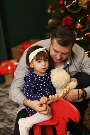 A young child in a navy blue polka dot dress and white headband holds onto a red rocking horse. A man embraces the child, holding a stuffed teddy bear. In the background, a Christmas tree decorated with ornaments and lights is visible, along with wrapped gifts.