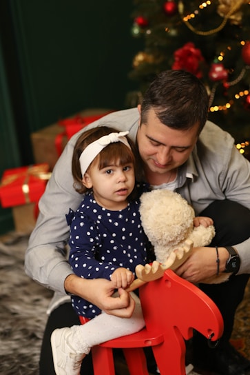 A young child in a navy blue polka dot dress and white headband holds onto a red rocking horse. A man embraces the child, holding a stuffed teddy bear. In the background, a Christmas tree decorated with ornaments and lights is visible, along with wrapped gifts.