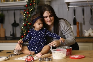 A woman and a young child are in a kitchen setting, engaged in baking activities. The child, sitting on a wooden table, is reaching towards a container with flour. Various baking tools and utensils are visible, including cookie cutters, a rolling pin, and a toy car. In the background, there is a decorated Christmas tree with lights, adding a festive atmosphere to the scene.