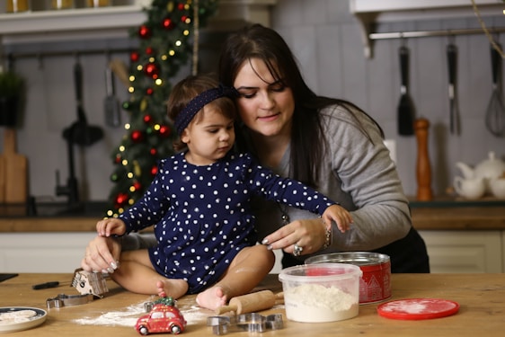 A woman and a young child are in a kitchen setting, engaged in baking activities. The child, sitting on a wooden table, is reaching towards a container with flour. Various baking tools and utensils are visible, including cookie cutters, a rolling pin, and a toy car. In the background, there is a decorated Christmas tree with lights, adding a festive atmosphere to the scene.