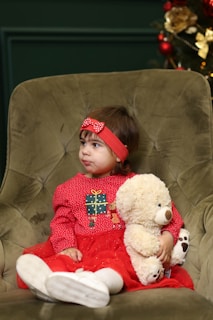 A young child wearing a red dress with a decorative gift box design on the front, sitting on a green armchair. The child has a red headband with a bow and holds a fluffy white teddy bear. In the background, there is a partially visible Christmas tree adorned with ornaments.