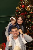 A family poses together in front of a decorated Christmas tree. The father is sitting with his daughter on his shoulders, while the mother stands beside them, smiling. The tree is adorned with baubles, lights, and flowers, creating a festive atmosphere.